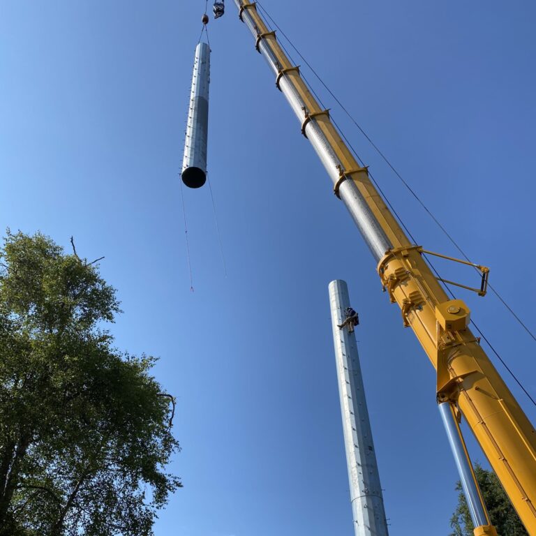 A yellow crane takes long, circular pieces of a tower into the air.